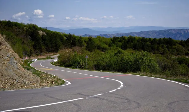 Çam ağaçları ve yeşil çalılar arasında virajlı, boş bir dağ yolu, uzakta sıra sıra dağlar