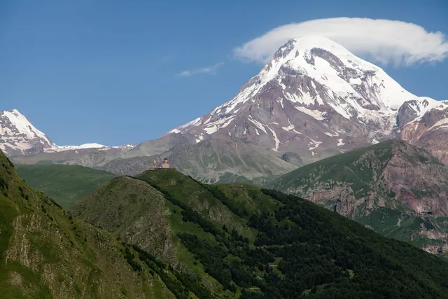 Yeşil bir sırtta minicik görünen Gergeti Kilisesi, üzerinde zirvesinde mercek biçimli bulut olan Kazbek