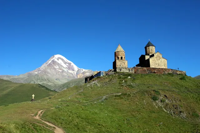 Gergeti Kilisesi'nin altındaki yeşil yamaçta toprak patika ve taş haç, arkada Kazbek